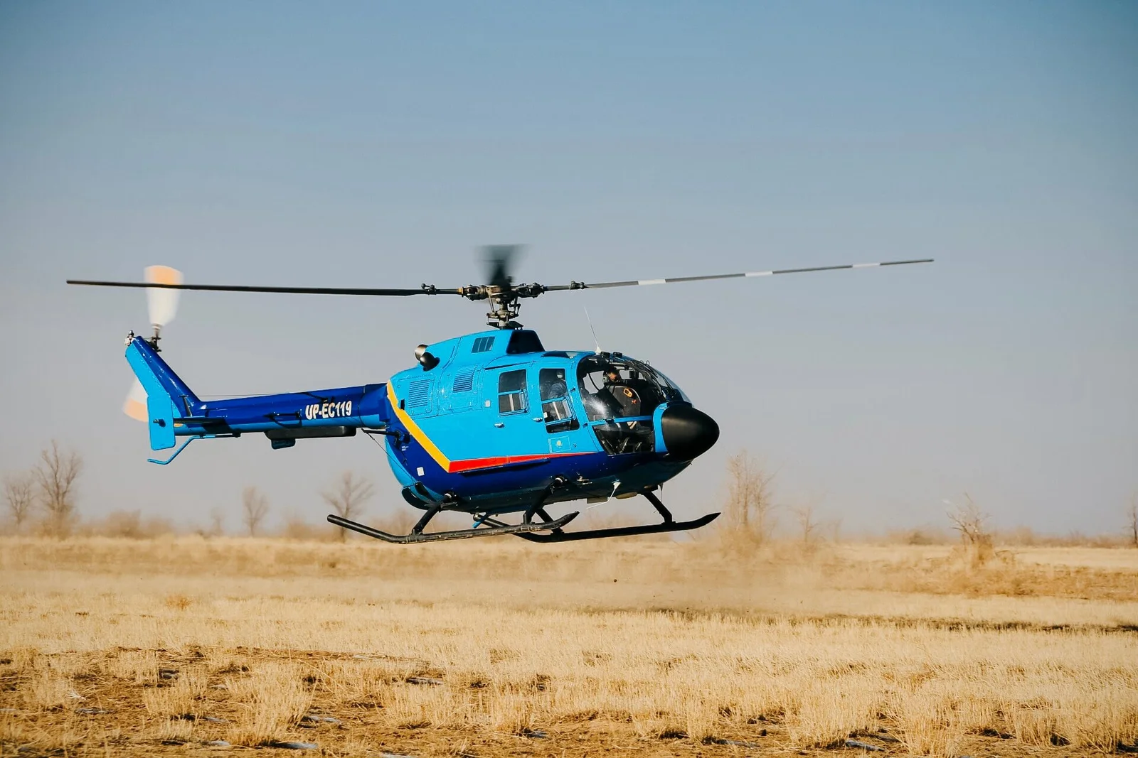Blue helicopter taking off low over a dry grass field near Almaty